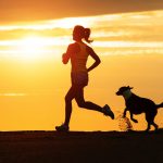 Woman And Dog Running On Beach At Sunset